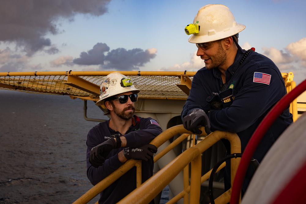 Two offshore workers in safety gear talking on an oil platform at sunset.