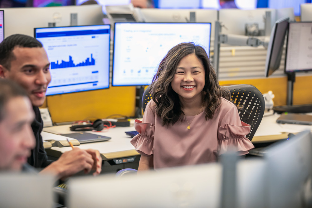 bp employees in an office trading environment. A woman in a pink blouse is smiling while seated at her desk, with charts displayed on computer monitors in the background.