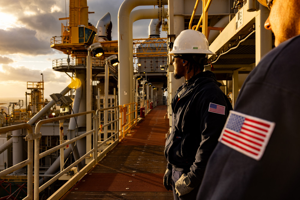 Engineers in protective gear with American flag patches standing on an oil and gas platform at sunset.